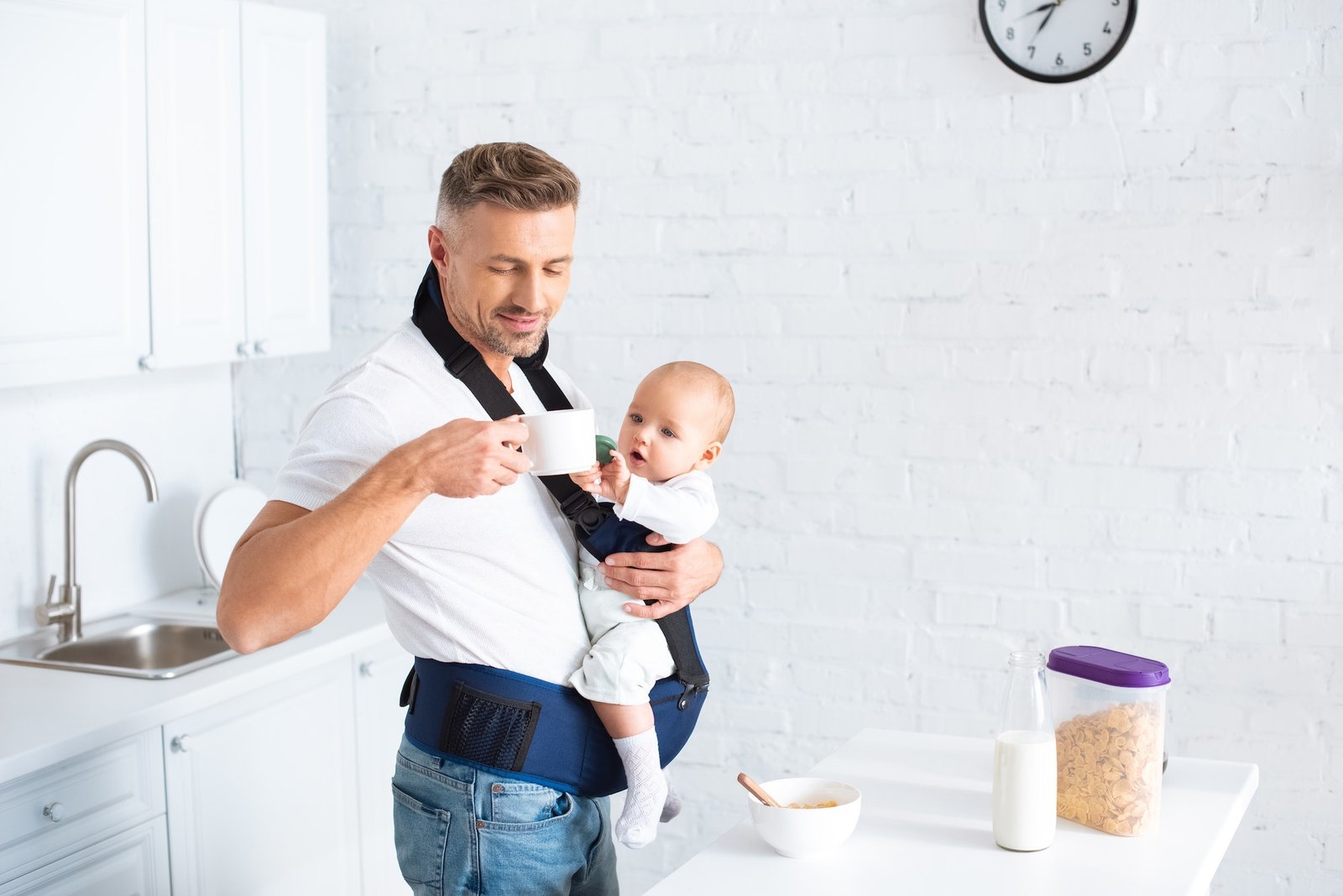 father holding infant daughter in baby carrier and looking at cup in kitchen